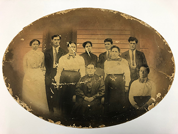 Horizontally-oriented oval group portrait of the Brown Family, 1912. Six women and three men are posed against a building's exterior wall.