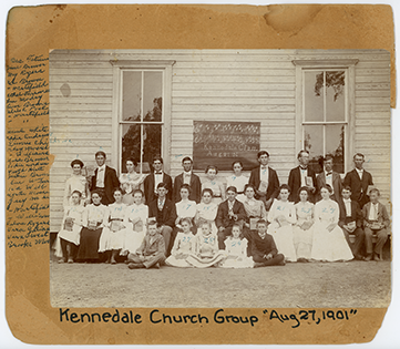 Group portrait of the Kennedale First Baptist Singing School posed against the exterior wall of a building. Blackboard on wall behind group with a musical score and words Kennedale Class, August 27, '01. Handwritten inscription below the image reads Kennedale Church Group August 27, 1901.